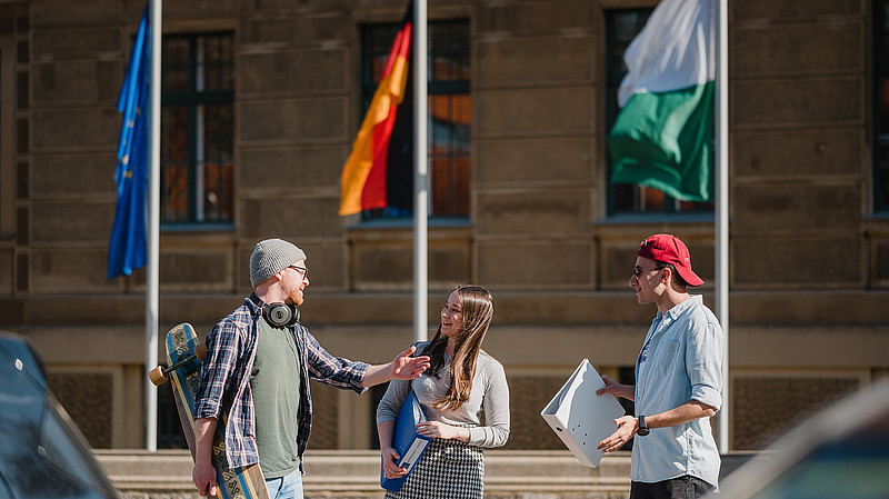 Internationale Wirtschaftskommunikation studieren in Görlitz. Studentische Arbeitsgruppe sitzt am Tisch vor einer Visualisierung. Drei Studierende vor einem Gebäude. Im Hintergrund Fahnen.