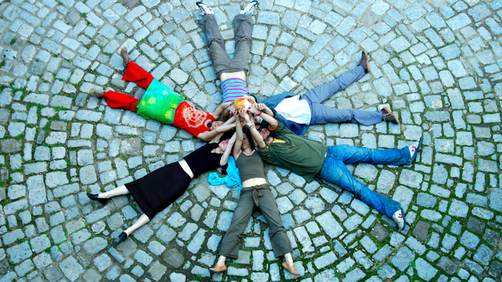 Auf einem Steinboden liegen Studierende im Kreis und strecken sich die Arme entgegen zu einem Stern.