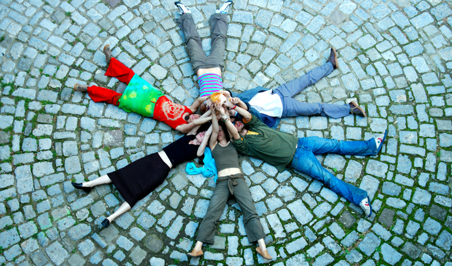 Auf einem Steinboden liegen Studierende im Kreis und strecken sich die Arme entgegen zu einem Stern.