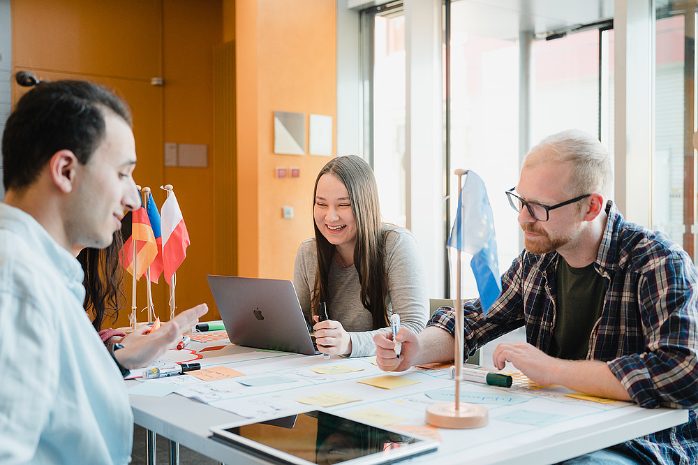 Studierende sitzen am Schreibtisch und arbeiten an einem Projekt. Vor ihnen liegt ein Plakat mit Klebezetteln, daneben liegen Tablets und Notebooks. Auf dem Tisch stehen die deutsche, polnische und tschechische Flagge.