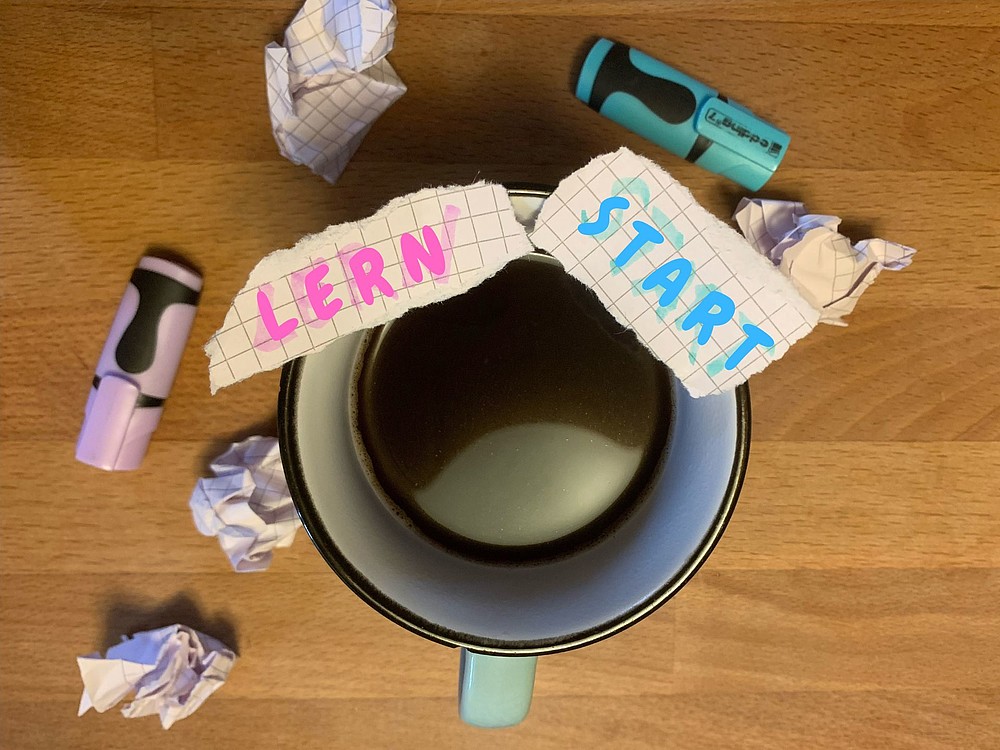 Blick von oben auf eine gefüllte Kaffeetase mit Papierstücken drauf, beschriftet mit den Worten Lern und Start.